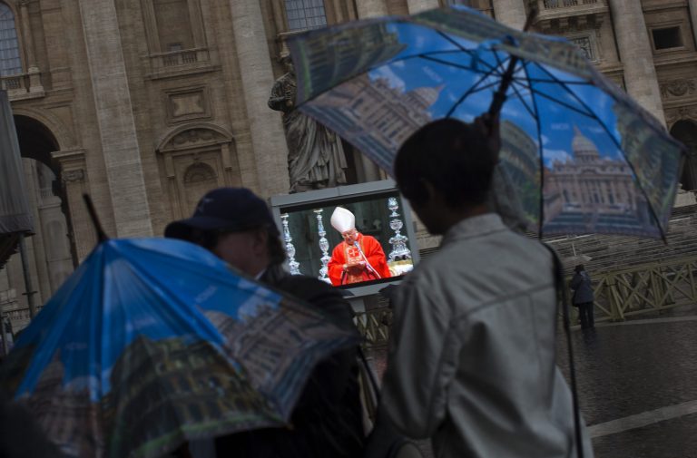 Faithful protect themselves from the rain as they follow a mass inside St. Peter's Basilica celebrated by Cardinal Angelo Sodano, seen on a giant screen in St. Peter's Square, Tuesday, March 12, 2013. Cardinals enter the Sistine Chapel on Tuesday to elect the next pope amid more upheaval and uncertainty than the Catholic Church has seen in decades: There's no front-runner, no indication how long voting will last and no sense that a single man has what it takes to fix the many problems. (AP Photo/Emilio Morenatti)