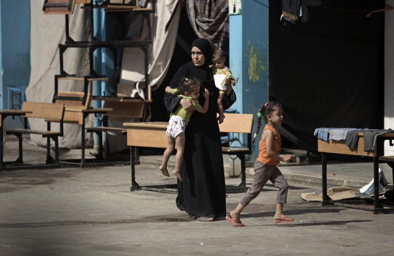In this Wednesday, Sept. 3, 2014 photo, a Palestinian woman carries two children as she walks in a U.N. school where she lives with her family after her house was destroyed by Israeli strikes in Gaza City. Israeli airstrikes have left much of the territory in ruins, and thousands of homes have been destroyed or severely damaged. Reconstruction has yet to begin as a blockade imposed by Egypt and Israel on Gaza still holds, severely restricting the import of cement and other building materials. (AP Photo/Khalil Hamra)