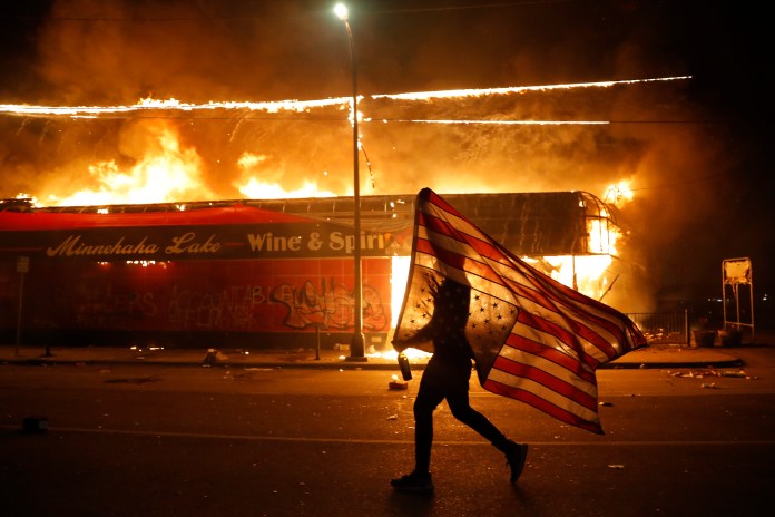 A protester carries a U.S. flag upside down, a sign of distress, next to a burning building, Thursday, May 28, 2020, in Minneapolis during protests over the death of George Floyd.