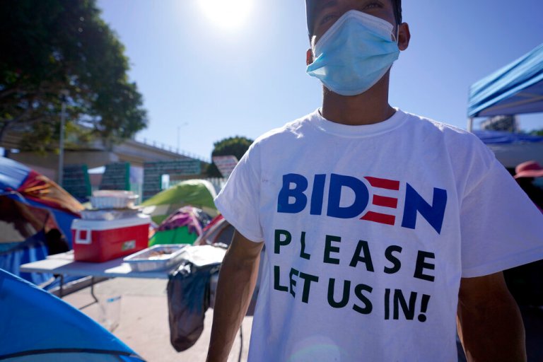 A Honduran man seeking asylum in the United States wears a shirt that reads, "Biden please let us in," as he stands among tents that line an entrance to the border crossing in Tijuana, Mexico.