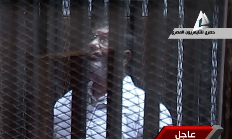 In this image taken from Egypt State TV,  Egypt's toppled President Mohammed Morsi stands inside a glass-encased metal cage in a courtroom in Cairo, Egypt, Tuesday, Jan. 28. 2014. Morsi was, separated from other defendants for the start of a new trial Tuesday over charges from prison breaks during the country's 2011 revolution, state television reported. (AP Photo/Egyptian State TV)