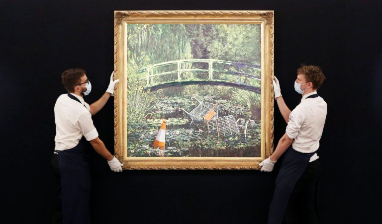 Two technicians handle Banksy's artwork,  'Show me the Monet' on display at Sotheby's in central London, Friday, Sept. 18, 2020. Banksy's contemporary re-interpretation of Claude Monet's depiction of the Japanese bridge in his garden at Giverny will be part of Sotheby's live-streamed auction on Oct. 21. (Jonathan Brady/PA via AP)