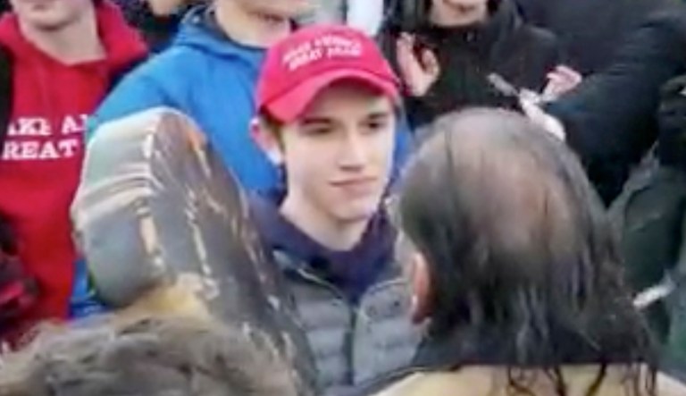 In this Friday, Jan. 18, 2019 image made from video provided by the Survival Media Agency, a teenager wearing a "Make America Great Again" hat, center left, stands in front of an elderly Native American singing and playing a drum in Washington. The Roman Catholic Diocese of Covington in Kentucky is looking into this and other videos that show youths, possibly from the diocese's all-male Covington Catholic High School, mocking Native Americans at a rally in Washington.