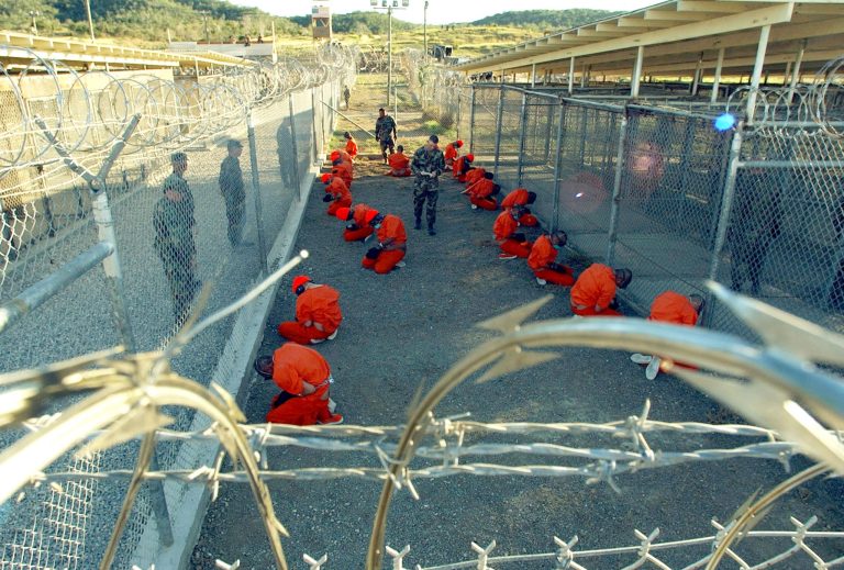 U.S. Military Police guard Taliban and al Qaeda detainees in orange jumpsuits January 11, 2002 in a holding area at Camp X-Ray at Naval Base Guantanamo Bay, Cuba during in-processing to the temporary detention facility. (Photo by Petty Officer 1st class Shane T. McCoy/U.S. Navy/Getty images)