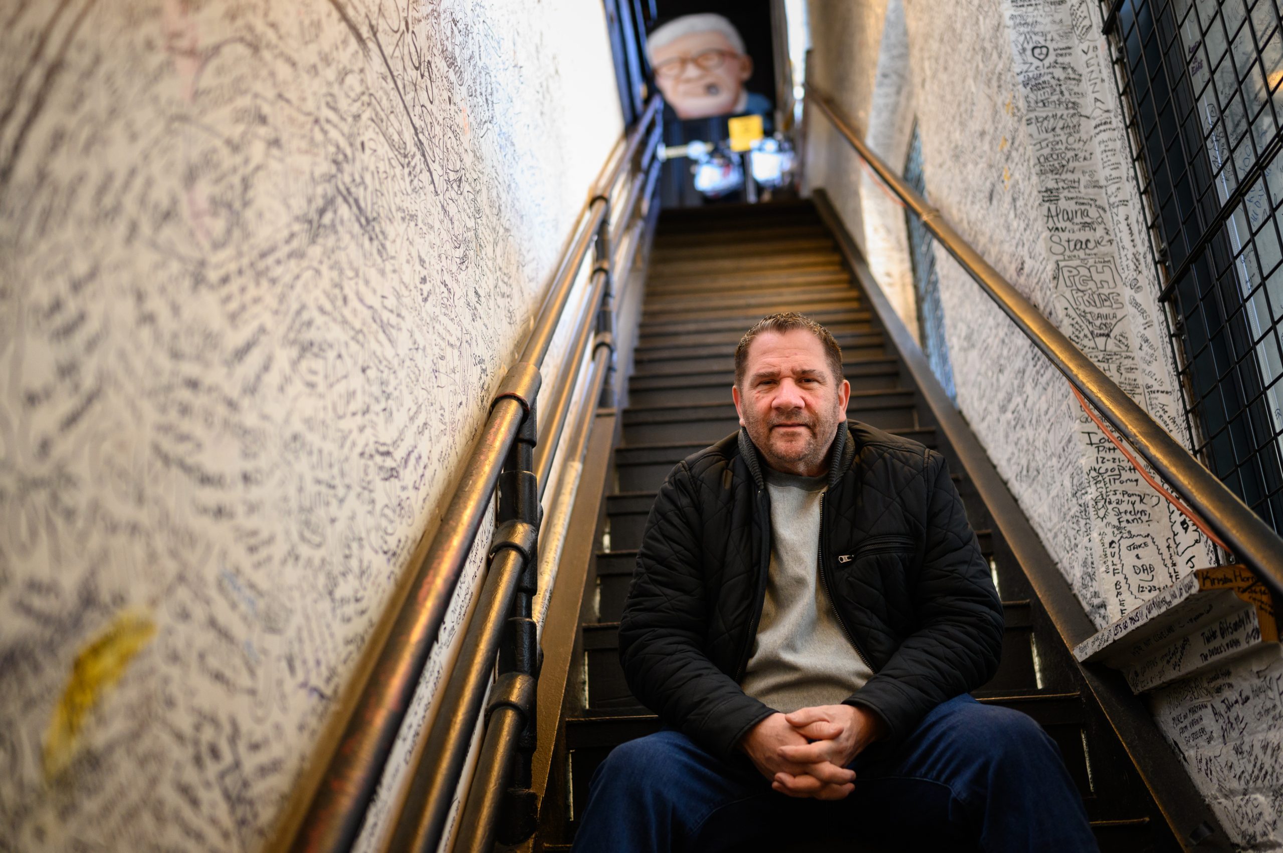 Jimmy Coen, owner of Yinzers in the Burgh, sits in a stairway signed by visitors to the store on Friday in Pittsburgh’s Strip District.↵↵