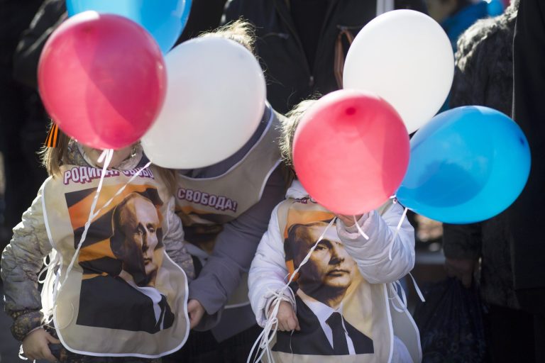 Two girls wearing tops  with a portrait of Russian President Vladimir Putin hold baloons in colors of Russian national flag stand in front of patriotic demonstrators gathered to support Russians in Crimea in Moscow, Russia, Monday, March 10, 2014. In Ukraine's Crimean peninsula a referendum has been called to see whether the region should split off and seek to become part of Russia is expected to held on Sunday . (AP Photo/Alexander Zemlianichenko)