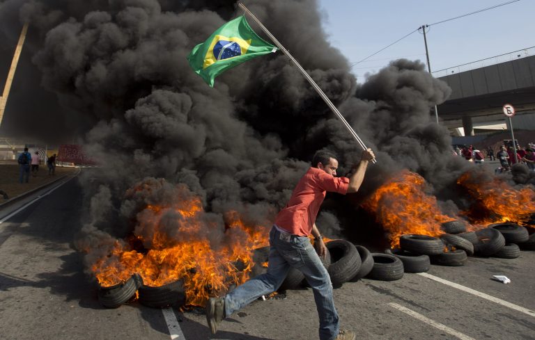 A member of the Homeless Workers Movement carries a Brazilian flag past burning tires during a protest against money spent on the World Cup near Itaquerao stadium which will host the international soccer tournament's first match in Sao Paulo, Brazil, Thursday, May 15, 2014. Brazilians are angry at the billions spent to host the World Cup, much of it on 12 ornate football stadiums, one-third of which critics say will see little use after the big event. (AP Photo/Andre Penner)
