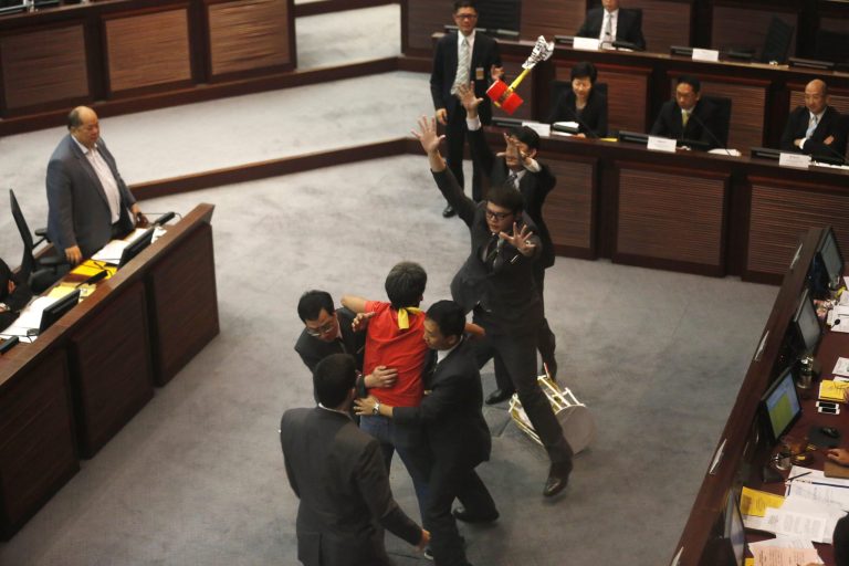 Pro-democracy lawmaker Leung Kwok-hung, wearing a red T-shirt in center, is stopped by security guards as he throws a plastic hammer towards Hong Kong Chief Secretary Carrie Lam, third from right,  during a meeting on proposing electoral reforms at the Legislative Council in Hong Kong Tuesday, July 15, 2014. The chief executive has submitted a report to the National People's Congress Standing Committee on political reform, saying that the majority of Hong Kong people are eager to have universal suffrage in the chief executive election in 2017.  (AP Photo/Kin Cheung)