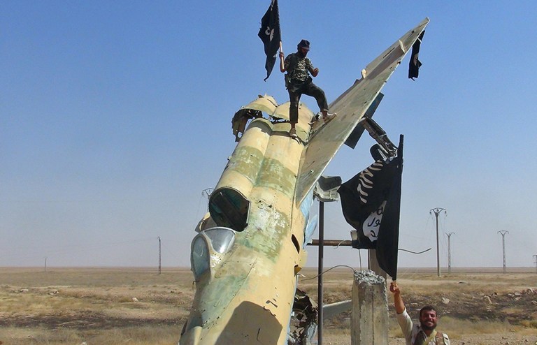 FILE - This undated image posted on Aug. 27, 2014 by the Raqqa Media Center of the Islamic State group, a Syrian opposition group, which has been verified and is consistent with other AP reporting, shows fighters of the Islamic State group waving the group's flag from a damaged display of a government fighter jet following the battle for the Tabqa air base, in Raqqa, Syria.   The Islamic State group is test flying, with the help of former Iraqi air force pilots, several fighter jets captured earlier from air bases belonging to the Syrian military, a Syrian activist group said Friday, Oct. 17, 2014. The report by the Britain-based Syrian Observatory for Human Rights could not be independently confirmed, and U.S. official said they had no reports of Islamic State group militants flying jets in support of their forces on the ground.(AP Photo/ Raqqa Media Center of the Islamic State group)
