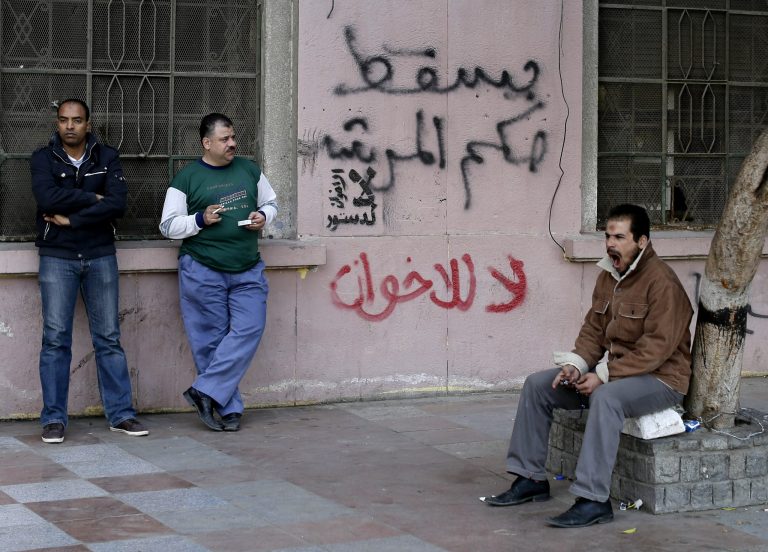   Egyptian men stand near Arabic writing on a wall in Arabic that reads, 