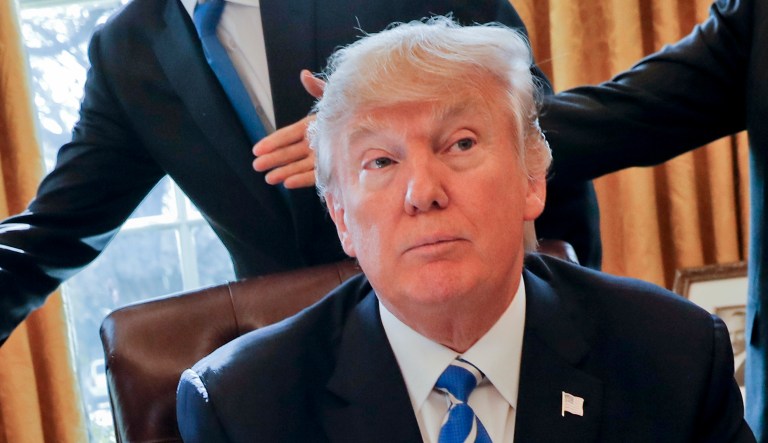 President Trump sits at his desk while others move around behind him.