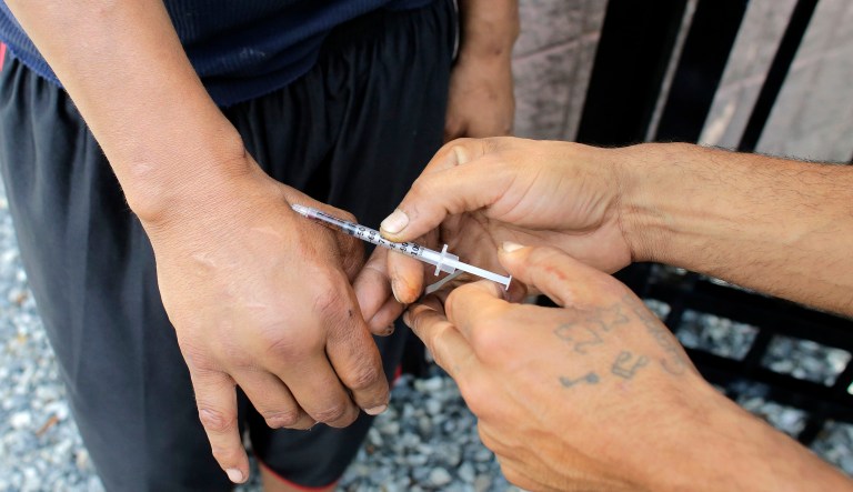 In this Wednesday, Aug. 23, 2017 photo an unidentified heroin user, left, is injected by another man, right, on the street near a strip of land sometimes referred to as "Methadone Mile," in Boston. Residents and those recovering from drug addiction say the area has seen little to no improvement after city officials promised new measures to tackle the notorious drug haven last summer. 