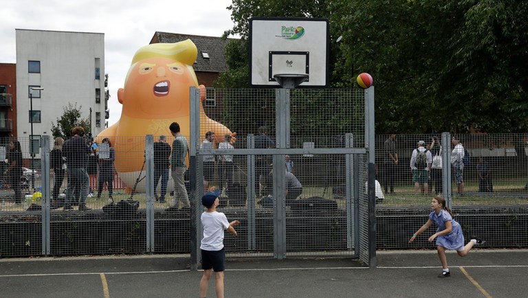 Children play basketball in the foreground as a six-meter high cartoon baby blimp of President Trump stands inflated during a practice session.