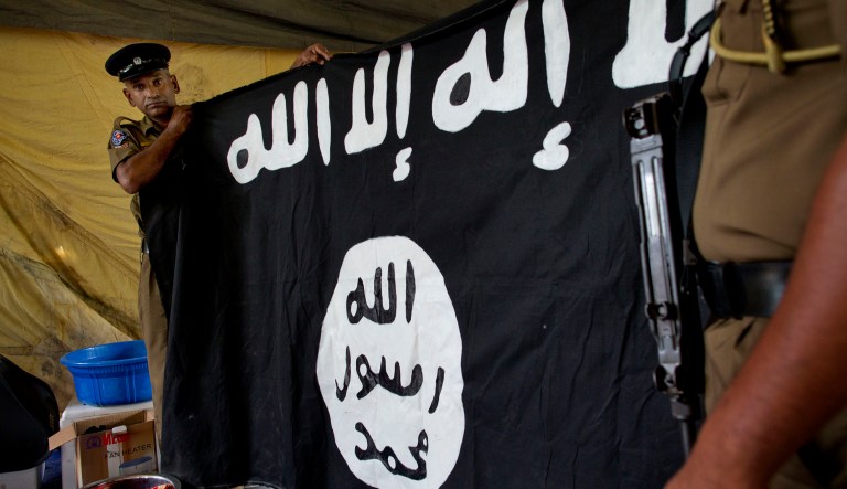 Police officers display a flag in Arabic that reads: "There is no god, but Allah" and "Of Allah is the Prophet, Muhammad" in Ampara, Sri Lanka, Sunday, April 28, 2019. Police in Ampara showed The Associated Press on Sunday the explosives, chemicals and Islamic State flag they recovered from the site of one security force raid in the region as Sri Lanka's Catholics celebrated at televised Mass in the safety of their homes.
