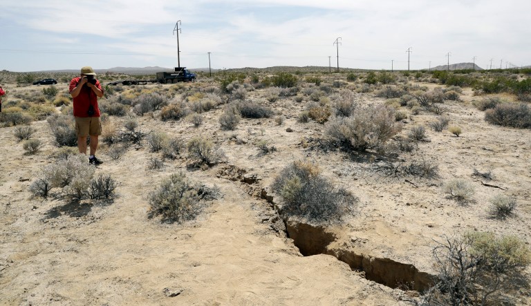 In this July 7, 2019 photo, a visitor takes a photo of a crack in the ground following recent earthquakes near Ridgecrest, Calif. The Naval Air Weapons Station China Lake military base nearby remains closed to non-essential personnel as damage assessments continue following last week's powerful earthquakes in the area. 