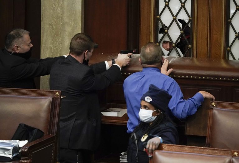 Police with guns drawn watch as protesters try to break into the House Chamber doors blocked by desk once used by pages.