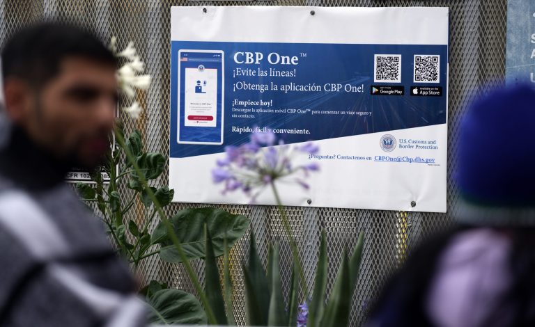 People waiting to apply for asylum stand in front of a sign for the CBP One app as they camp near the pedestrian entrance to the San Isidro Port of Entry, linking Tijuana, Mexico, with San Diego, Thursday, June 1, 2023, in Tijuana, Mexico. U.S. authorities raised the number of people allowed to enter the country with an online app.