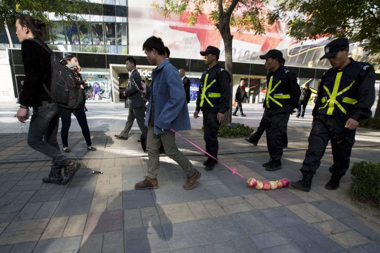 Chinese performance artist Han Bing, left, and his friend Hui Li, center, drag an iPhone and a string of apples, respectively, as they are escorted by security guards in a shopping district where an Apple retail store is located in Beijing, China, Thursday, Oct. 16, 2014. Han Bing, known to take a cabbage out on a walk to raise questions about our attachments to things, hopes to highlight our modern dependence on the mobile phone in this performance.  (AP Photo/Ng Han Guan)