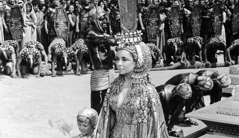 British actress Elizabeth Taylor in one of her most arduous and majestic scenes as âCleopatraâ, a 20th century-Fox spectacle filmed in Romeâs Cinecitta studios, Italy on May 8, 1962, portraying the Queen of Egyptâs momentous entry into Rome with young Caesarion (4-year-old Italian Loris Loddi) at her side. Nubian slaves bow as the âQueenâ steps off the platform while crowds throng behind a line of Roman legion soldiers for the most spectacular scene filmed on May 8, 1962. Miss Taylor is wearing an elaborate and gilded costume with sleeves shaped like wings. On her head is an ornamented head-gear studded with hard stones and which reaches her shoulders while her crown is lined with images of the Egyptian cobras supporting the sun. Topping the crown is the symbol of her Egyptian queenship. The entire head-gear weighs about six kilos.