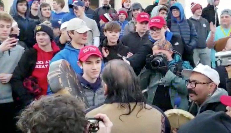 In this Friday, Jan. 18, 2019 image made from video provided by the Survival Media Agency, a teenager wearing a "Make America Great Again" hat, center left, stands in front of an elderly Native American singing and playing a drum in Washington. The Roman Catholic Diocese of Covington in Kentucky is looking into this and other videos that show youths, possibly from the diocese's all-male Covington Catholic High School, mocking Native Americans at a rally in Washington.