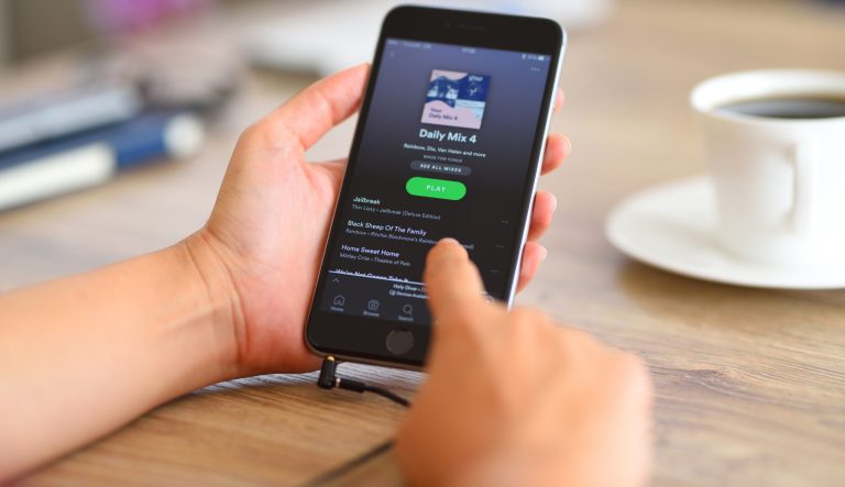 Woman using smart phone on a wooden desk. The smart phone is an iPhone 6 plus displaying Spotify app. 