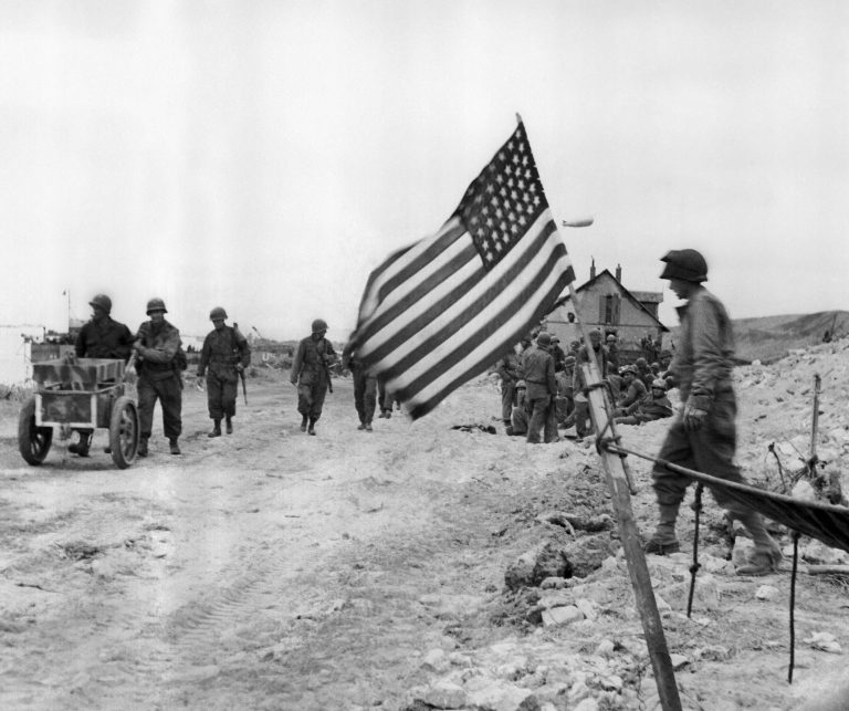 The American flag flies from a make-shift staff on a beach on the French Normandy coast on June 14, 1944 where allied troops fought bitterly to push back the Germans. Americans in background push a captured German ammunition cart. (AP Photo)