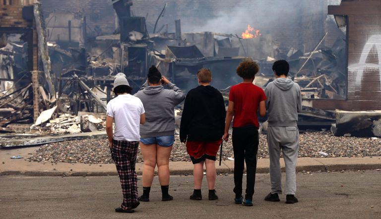 Onlookers watch as smoke smolders from a destroyed fast food restaurant near the Minneapolis Police Third Precinct, Thursday, May 28, 2020, after a night of rioting and looting as protests continue over the death of George Floyd, who died in police custody Monday night in Minneapolis after video shared online by a bystander showed a white officer kneeling on his neck during his arrest as he pleaded that he couldn't breathe. 