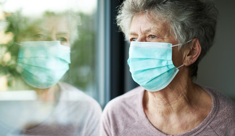 An elderly woman wearing a mask and looking out a window.
