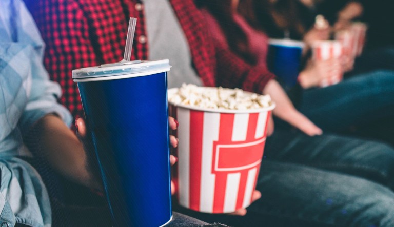 Close up of tasty but unhealthy food. There are basket of popcorn and a blue cup of coke on picture. Man and woman are holding it together. 