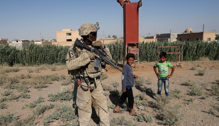 A U.S. soldier stands near Syrian children on a road that links to Raqqa, Syria, Wednesday, July 26, 2017. The U.S. has up to 1,000 troops in Syria mostly involved in training and advising the local forces against IS. Kurdish forces have gained confidence in light of open U.S. support to their forces, particularly as the battle for Raqqa took off. 