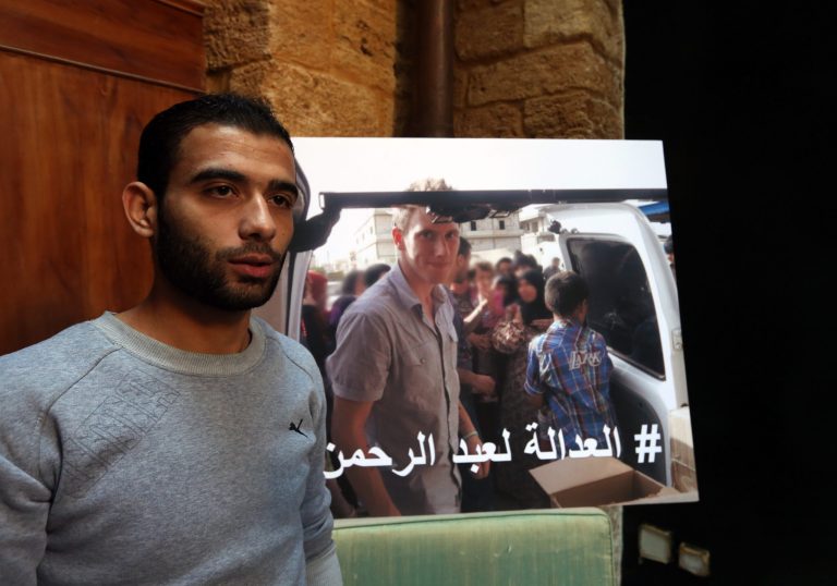 Syrian refugee Amjad Moghrabi stands in front of a photograph of his colleague, American aid worker Peter Kassig, 26, who converted to Islam while in captivity and changed his name to Abdul-Rahman Kassig, during an interview with The Associated Press in the northern port city of Tripoli, Lebanon, Saturday, Nov. 8, 2014. Kassig was helping victims of the Syrian civil war when he was captured in Syria last year and threatened with beheading by the Islamic State group. Arabic reads, 