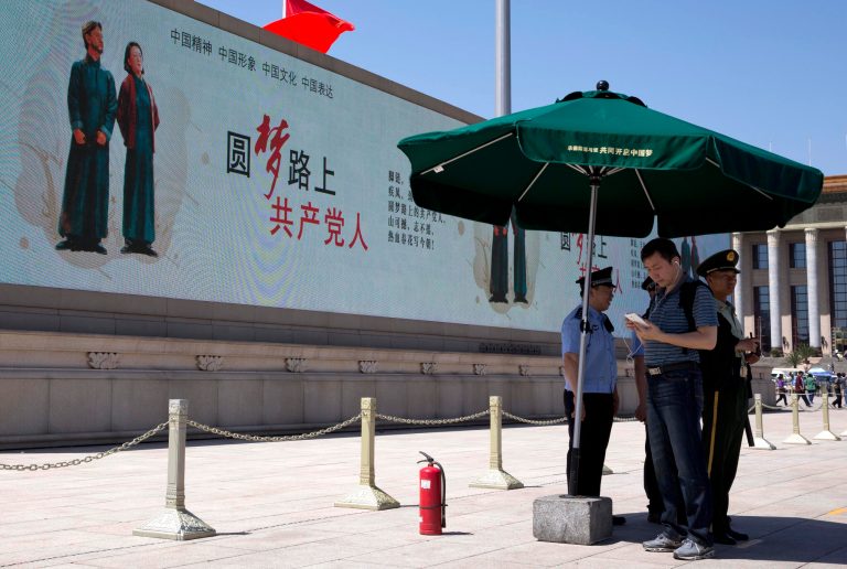 In this Wednesday, May 28, 2014 photo, a man looks at his smartphone next to Chinese policemen and a paramilitary policeman standing guard near a giant electronic screen showing a Chinese government propaganda message reading 