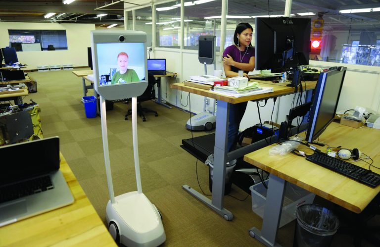 Senior software engineer, Josh Faust, seen on screen, navigates his company's office using a Beam remote presence system, as fellow engineer Stephanie Lee, at right, works on a project at Suitable Technologies in Palo Alto, Calif., Wednesday, Dec. 12, 2012. More employees are working from home, but there's still no substitute for actually being at the office. Enter the Beam. It's a roving computer screen _ with video cameras, microphones and speakers _ that stands five feet and rides on motorized wheels. (AP Photo/Marcio Jose Sanchez)