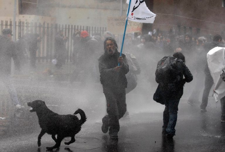   FILE - In this June 10, 2012 file photo, a dog runs in the spray as riot police use water cannons against demonstrators during a protest against the premiere of a documentary about the late Gen. Augusto Pinochet in Santiago, Chile. As the protests become fixtures in this modernizing capital, normally unnoticed street dogs have become stars in their own right. (AP Photo/Luis Hidalgo, File)  