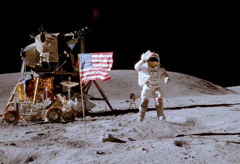 A spacesuit-clad Apollo astronaut stands proudly next to a red-white-and-blue American flag, his national trophy telling the lonely world 