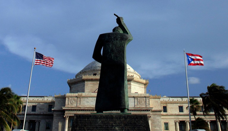 A bronze statue of San Juan Bautista stands in front of Puerto Rico's Capitol as U.S. and Puerto Rican flags fly in San Juan, Puerto Rico. (AP Photo/Ricardo Arduengo)