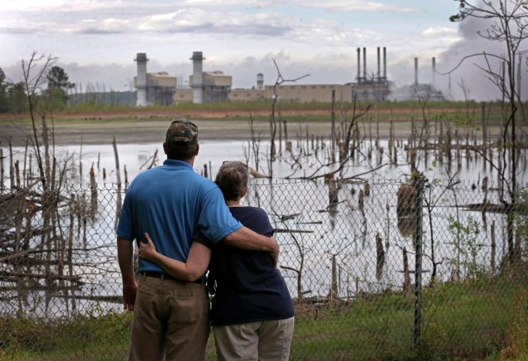 In this April 25, 2014 photo, Bryant Gobble, left, hugs his wife, Sherry Gobble, right, as they look from their yard across an ash pond full of dead trees toward Duke Energy's Buck Steam Station in Dukeville, N.C. Records obtained by The Associated Press show that Duke and North Carolina environmental regulators have known since 2011 that groundwater samples taken from monitoring wells near several homes in Dukeville contained substances, some that can be toxic, exceeding state standards. (AP Photo/Chuck Burton)