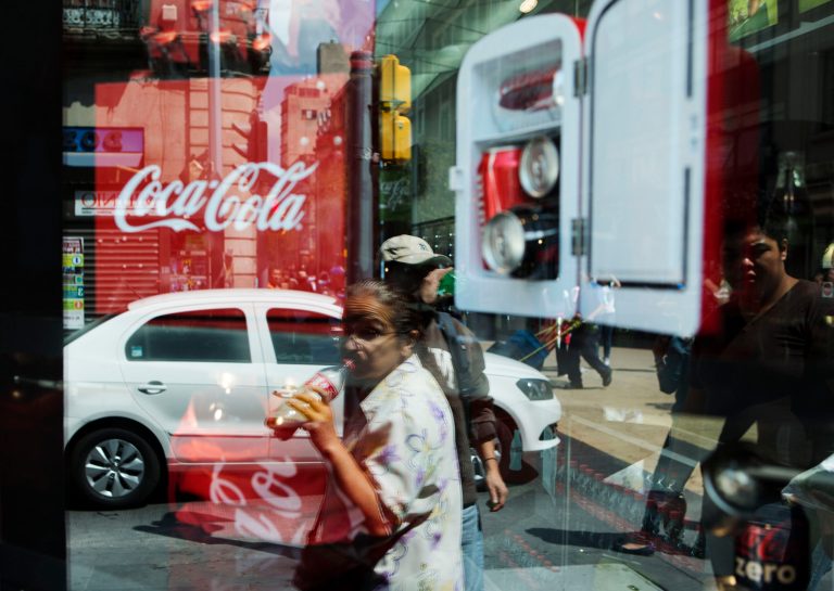A woman is reflected in a Coca-Cola store window display as she drinks a Coke in Mexico City, Thursday, Oct. 9, 2014. Coke and Pepsi are spending millions of dollars to oppose proposed new taxes on sugared drinks in California. Pepsico reported higher earnings Thursday, except in Mexico, where they blamed a drop in sales volume on a new tax on non-nutritional food and drinks. (AP Photo/Rebecca Blackwell)