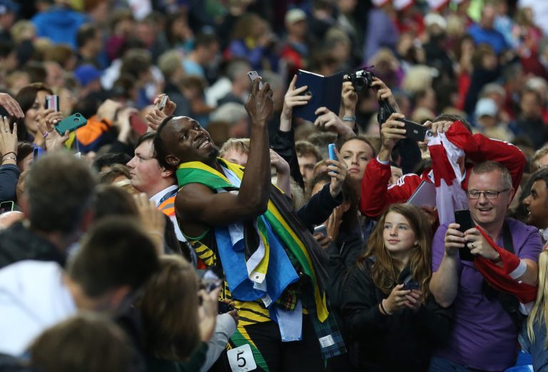 Usain Bolt of Jamaica takes a photo on a smart phone with a member of the crowd as he walks round Hampden Park Stadium after he competed in the men's 4x100 meter relay race during the Commonwealth Games 2014 in Glasgow, Scotland, Saturday Aug. 2, 2014. (AP Photo/ Scott Heppell)