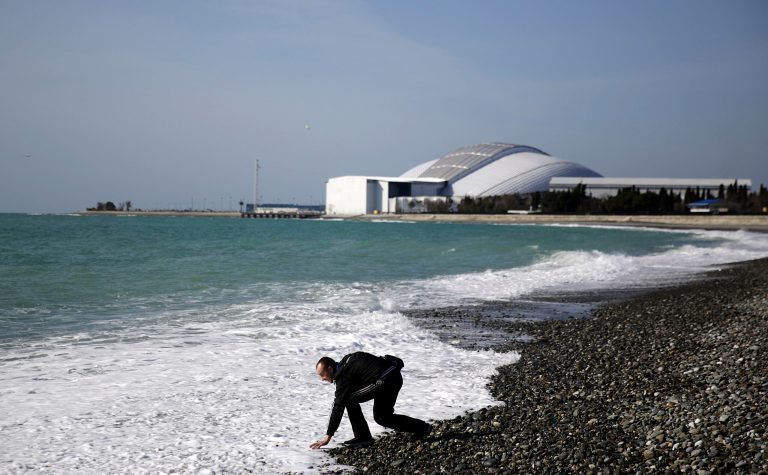 A beachgoer reaches down to touch the Black Sea as Fisht Olympic Stadium stands in the background, home of tonight's 2014 Winter Olympics closing ceremony, Sunday, Feb. 23, 2014, in Sochi, Russia. (AP Photo/David Goldman)