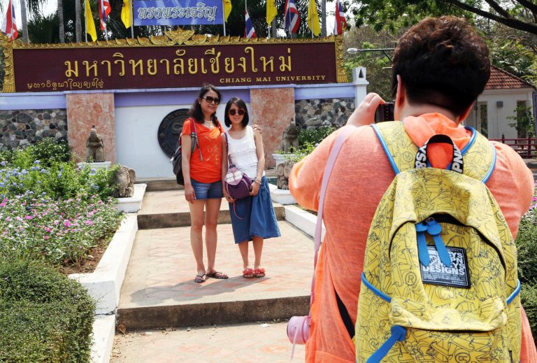 Advance for Thailand Challenging Chinese, in this photo taken March 30, 2014, Chinese tourists pose for photograph at the main entrance to Chiang Mai university in Chiang Mai province, northern Thailand. The bucolic, once laidback, campus of one of Thailandâs top universities seems to be under a security clampdown these days. Not against a terrorist threat, but Chinese tourists, thousands of them, who have clambered aboard student buses, eaten in cafeterias, sneaked into classes to attend lectures and even pitched a tent by a picturesque lake. (AP Photo/Apichart Weerawong)