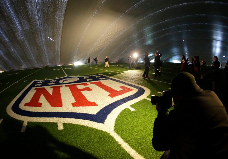 FILE - In this Jan. 15, 2014 file photo, the NFL logo at midfield of MetLife Stadium is illuminated by lights on television reporters' videocameras as members of the media are given a tour under a tarp used by crews to keep the turf dry ahead of Super Bowl XLVIII in East Rutherford, N.J. As the Denver Broncos and Seattle Seahawks prep for their Feb. 2, 2014 showdown in the Super Bowl, a legal fight is playing out over the very turf installed months ago at the NFL title game's venue. Taylor Turf Installation Inc. is suing the MetLife Stadium's operators and the company that hired the suburban St. Louis company, seeking more than $292,000 that Taylor Turf claims it still is owed for hustling to install the  stadium's playing surface last summer. (AP Photo/Julio Cortez, File)