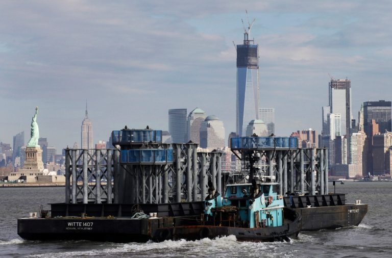   A barge loaded with sections of spire for One World Trade Center, center, is guided by tugboat across New York Harbor, Tuesday, Dec. 11, 2012 in New York. The Statue of Liberty is at left, and the Empire State Building is second left. (AP Photo/Mark Lennihan)  
