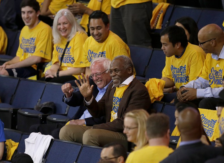 Democratic presidential candidate Sen. Bernie Sanders, I-Vt., center left, waves next to actor Danny Glover during Game 7 of the NBA basketball Western Conference finals between the Golden State Warriors and the Oklahoma City Thunder in Oakland, Calif. (AP Photo/Ben Margot)
