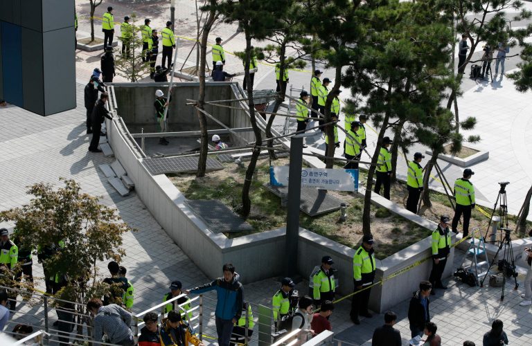 Police officers stand around a collapsed ventilation grate in Seongnam, South Korea, Saturday, Oct. 18, 2014. Sixteen people watching an outdoor pop concert in South Korea fell 20 meters (60 feet) to their deaths Friday when a ventilation grate they were standing on collapsed, officials said. (AP Photo/Lee Jin-man)
