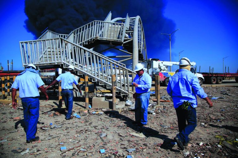 Rescue workers stand in front of damaged pedestrian bridge after a explosion in the Amuay refinery near Punto Fijo, Venezuela, Sunday, Aug. 26, 2012. Venezuelans who live next to the country's biggest oil refinery said they smelled a strong odor of sulfur hours before a gas leak ignited in an explosion on Saturday that killed at least 39 people and injured more than 80. (AP Photo/Ariana Cubillos)