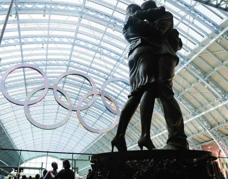 A couple pauses at the foot of a statue with the Olympic rings hung in the rafters of St. Pancras Station prior to the 2012 Summer Olympics, Tuesday, July 24, 2012, in London. The opening ceremonies for the 2012 London Olympics will be held Friday, July 27. (AP Photo/Charles Krupa)