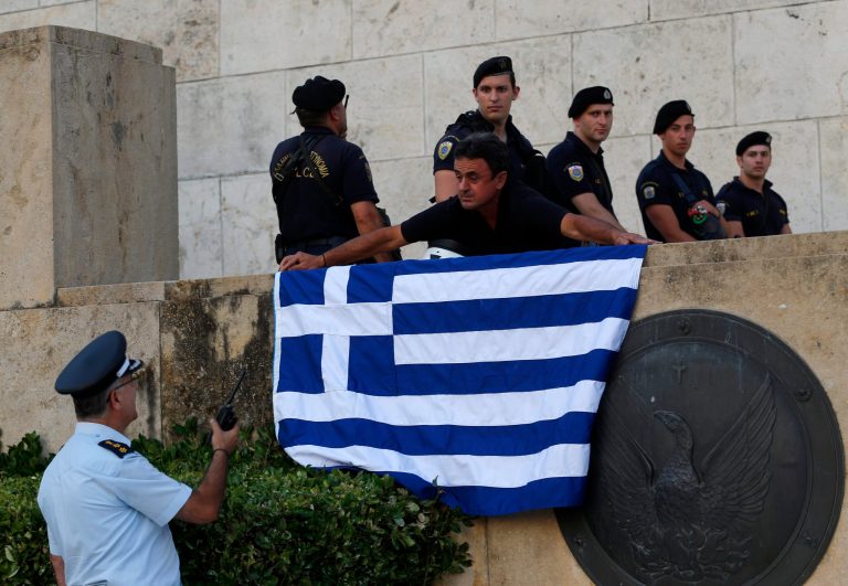 An anti-austerity protester places a Greek flag next to riot policemen as they guard one of the entrances of the Greek Parliament during a rally against the government's agreement with its creditors in Athens, Monday, July 13, 2015. After months of acrimony, Greece clinched a preliminary bailout agreement with its European creditors on Monday that will, if implemented, secure the country's place in the euro and help it avoid financial collapse. (AP Photo/Emilio Morenatti)