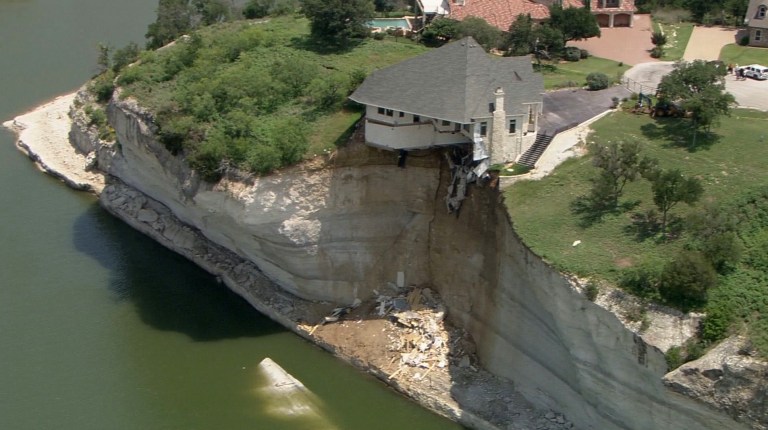 In this image taken from video provided June 12, 2014, by WFAA.Com, a luxury house teeters on a cliff about 75 feet above Lake Whitney in Whitney, Texas. The owner of a vacant luxury house teetering on a crumbling 75-foot cliff over a Central Texas lake has decided to burn the house and clear the lot of the debris. (AP Photo/WFAA.Com)