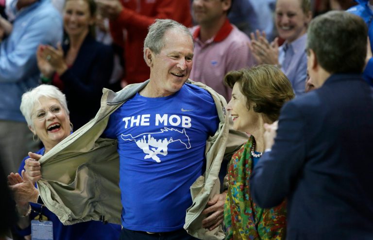 Former President George W. Bush opens his jacket to show off a t-shirt during the first half of an NCAA college basketball game between Cincinnati and SMU in Dallas, Sunday, Feb. 12, 2017. (AP Photo/LM Otero)